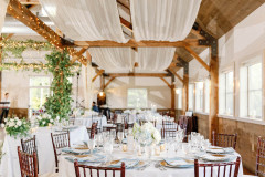 room with wooden rafters and white drapery with white round tables and wooden chairs.