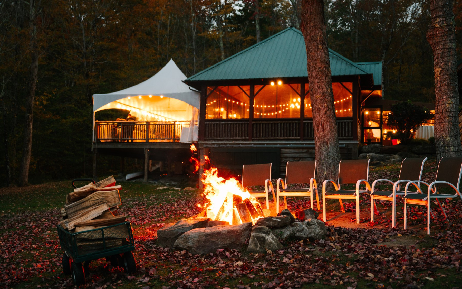 beach pavilion exterior featuring a white tent on the deck portion with a green roof on the main building. front of image showcases a firepit ablaze with a few chairs surrounding it and a wagon full of wood next to it.