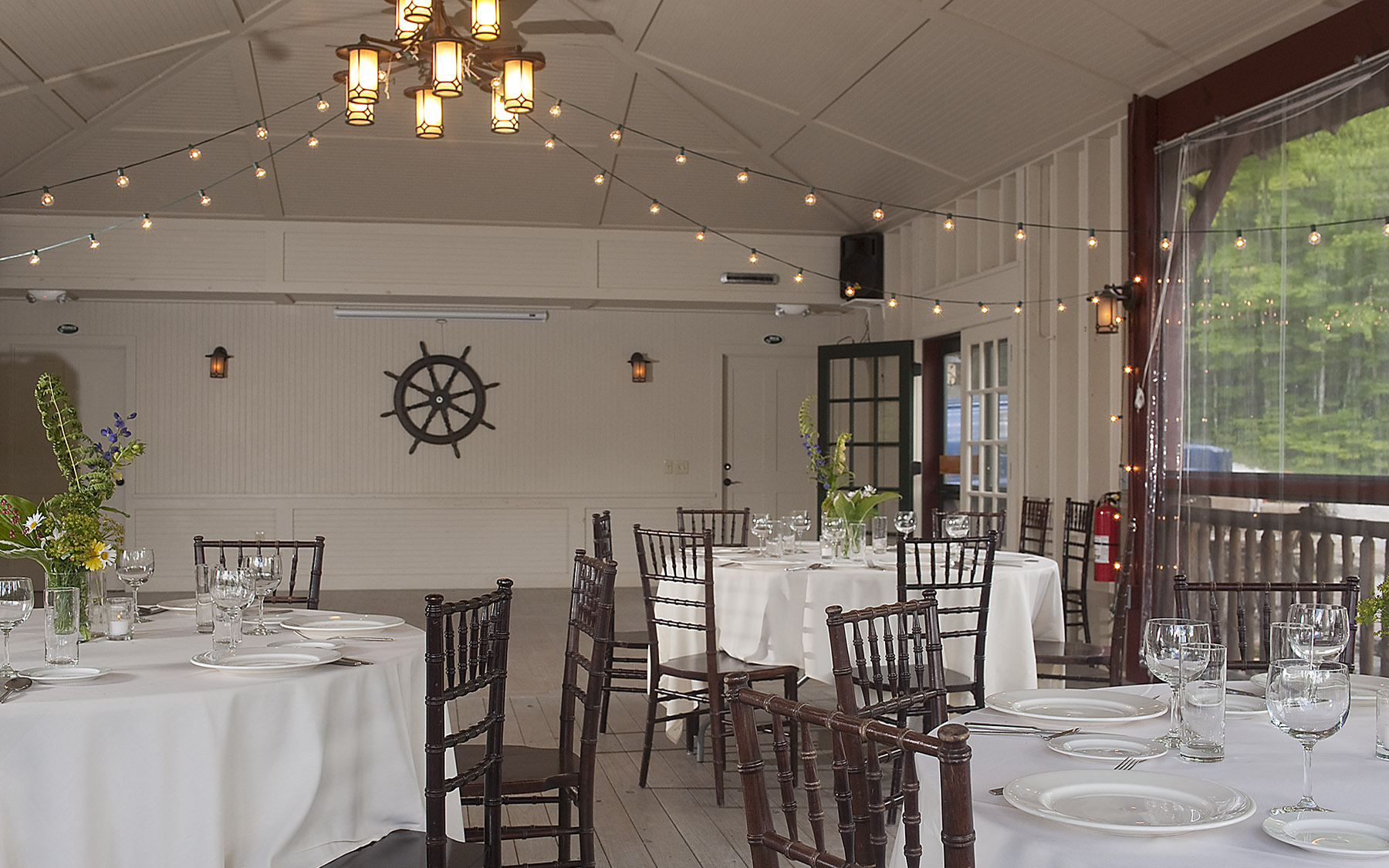 interior of room set for event with white table clothes, lanterns and a ships wheel on the wall.