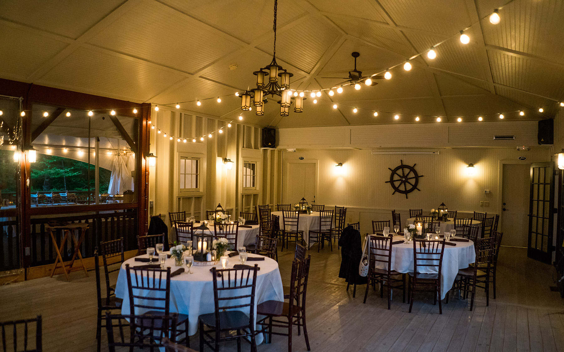 interior of room set for event with white table clothes, lanterns and a ships wheel on the wall.