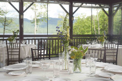 interior of room set for event with white table clothes, lanterns and view toward lake and woods through large windows.