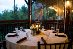 interior of room set for event with white table clothes, lanterns - view toward window facing lake and woods.