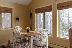 photograph of cloud nine guest home dining area. three windows on the walls have blinds lifted to reveal the view. table is white with a wooden top, and features four chairs around it.
