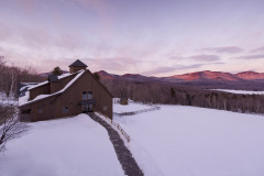 brown wooden barn in winter surrounded by snow with mountains in the background and violet sunset lighting.