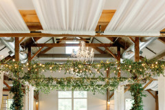interior of event barn set for wedding reception with white draperies, greenery, white table cloths and bamboo chairs.