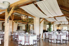 interior of event barn set for wedding reception with white draperies, greenery, white table cloths and bamboo chairs.