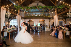 Event barn wedding reception with bride and groom dancing in center of room. lights, drapery and greens hanging from rafters. Guests sitting in chairs watching.