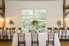 Room with wood floors and exposed beams and large window in center of white wall.  tables set with tall white flowers, white linens and wooden chairs.