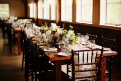 a row of wooden tables with wooden chairs and white flowers and tables set for dinner along a row of windows.