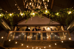 Wedding guests standing along railing looking down on lights and greens strung along rafters.