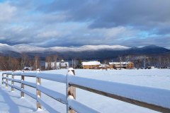 split rail fence along snow covered field with building and mountains in backround.