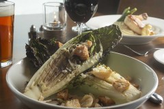 Grilled Caesar dish on table with glass of red wine and pint of amber beer.