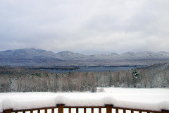 wooden railing covered in snow in the foreground with snowy trees, lake and mountains in the background with a cloudy sky.