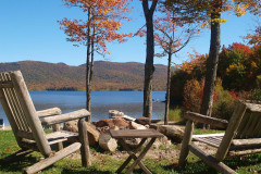 rustic timber chairs surrounding fire pit in front of lake surrounded by fall trees.