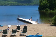 a metal dock on a lake with a sandy beach and lounge chairs. Children on dock about to jump into lake.