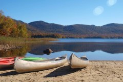 Canoes parked on a tan sand beach with a lake, mountains and blue sky in background.