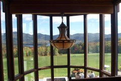 A view through a divided light window with a large stained glass chandelier hanging in the center. the outside view is of green grass and foliage covered mountains.