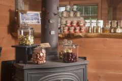 Vintange cast iron wood stove with glass jars sitting on it holding chocolates and multi colored mugs on a shelf with honey behind it.