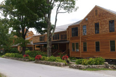 Exterior of tan wood sided building along road with stone wall and flowers in garden, green trees along front.