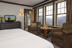 Bedroom with tv sitting on dark wood dresser, bed dressed in white, row of windows with view out to snowy mountains.