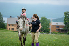 Small child in riding helmet on pony being led by woman in riding gear with wooden building and green  mountains in background.