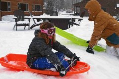 a young girl with brown hair, pink snow goggles and snow attire on an orange sleigh about to go down a hill. Another person in a tan quilted coat with a green sleigh is in the background.