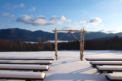 a birch arbor in the center of snow covered benches, snow covered meadow and trees, lake and mountains in the background with blue sky