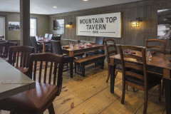 Large white wooden sign on wall reading Mountain Top Tavern 1940 in a room with wooden tables and chairs and wide plank wood flooring.