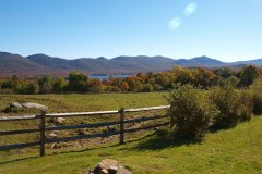 A view of a field with a split rail fence, green grass and foliage trees and mountains in background. Abundant blue sky.