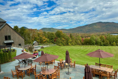 Outdoor trrace with tables and guests dining, grassy lawn and mountains in background.