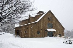 wooden post and beam barn in snow with snow covered tree to left, split rail fence to right and wreath hung on upper peak.