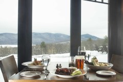 View across table with plate of wings, bone-in chicken dish and grilled caesar through windows to snowy mountains.