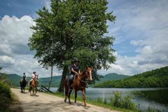 3 people riding 3 horses along a lake with summer trees and mountains in background.