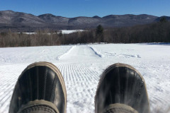 the tips of 2 winter boots in the foreground as though sitting on a sled at the edge of a groomed snowy trail with woods and mountains in the background.