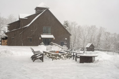 Mountain Top Resort adirondak chairs around a fire pit covered in snow with brown barn in backgfround.