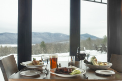 wooden table set with white plates, red wine and pint of amber beer. view out windows toward snowy mountains.