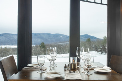 wooden table set with empty white plates and glassware with view out windows toward snowy mountains.