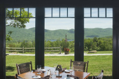 view of wooden table with empty place settings by large windows with views out to green mountains and blue sky in summer.