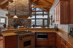 Mountain Top Resort Jewel Guest Home view of kitchen from entryway featuring stove and microwwave, large fireplace in the middle fo the room, and a view of snowy mountains in the background through large floor to ceiling windows.