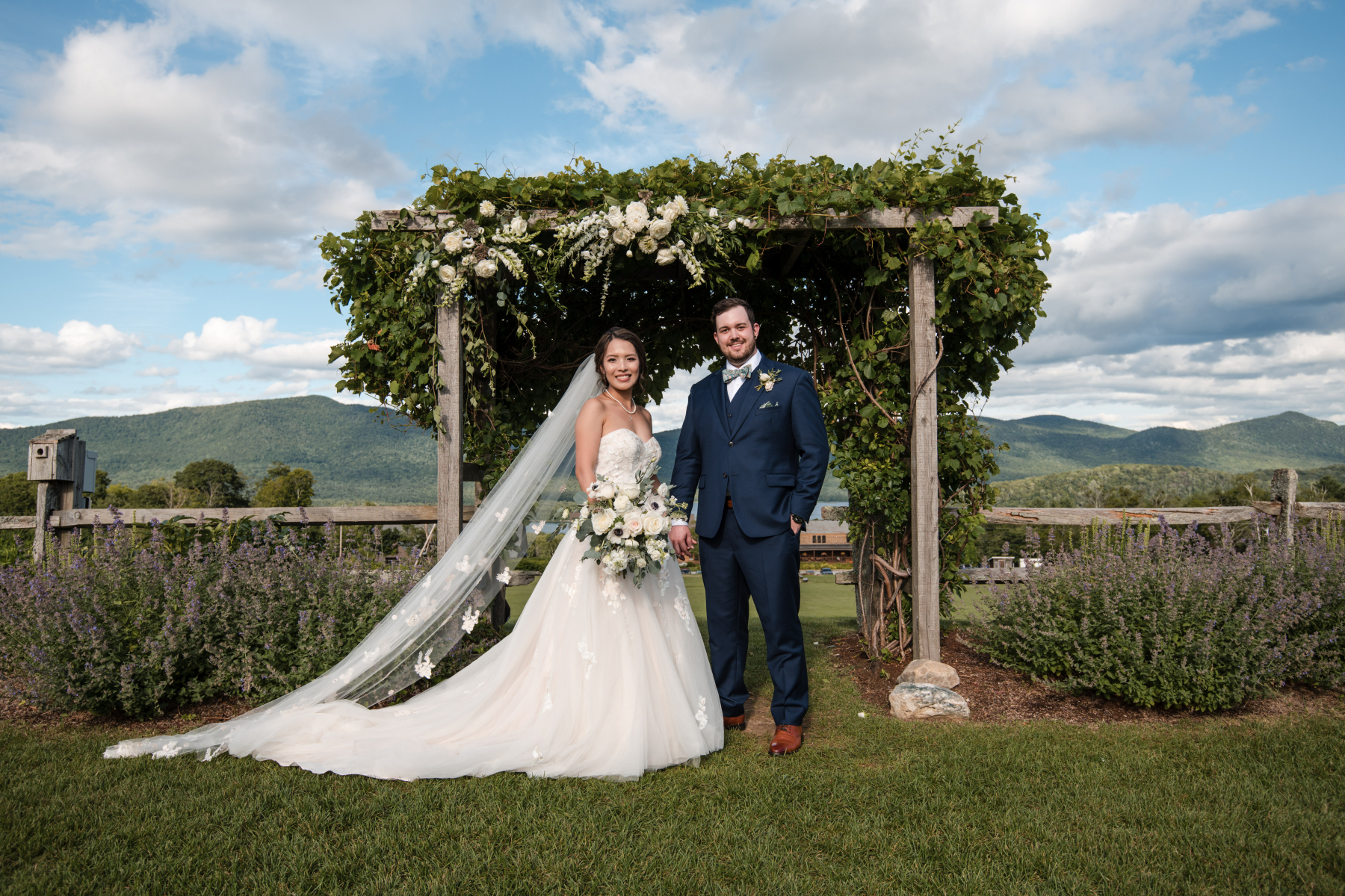 Bride in white, groom in dark suit, standing in front of arbor with greenery and white flowers.