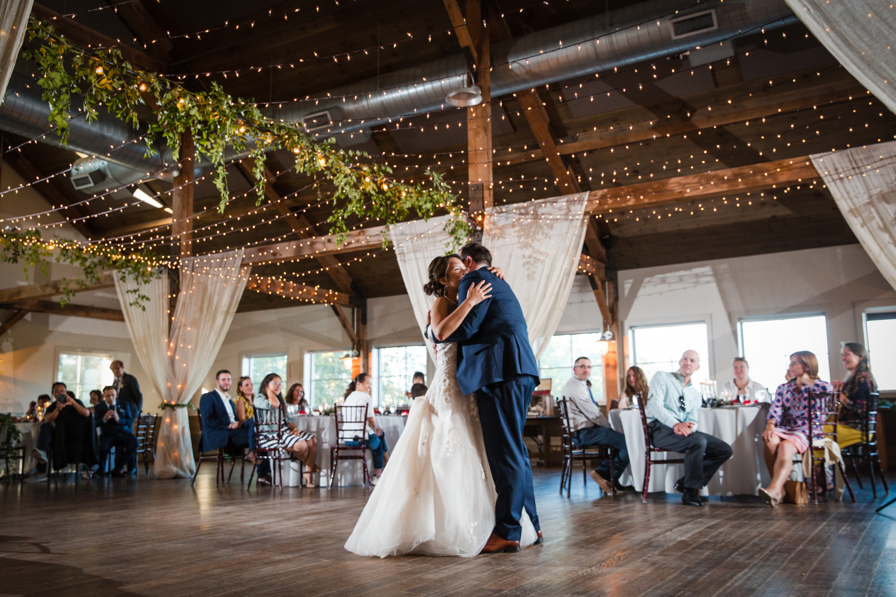 Bride and groom in traditional wedding attire having their first dance as guests in event barn look on.
