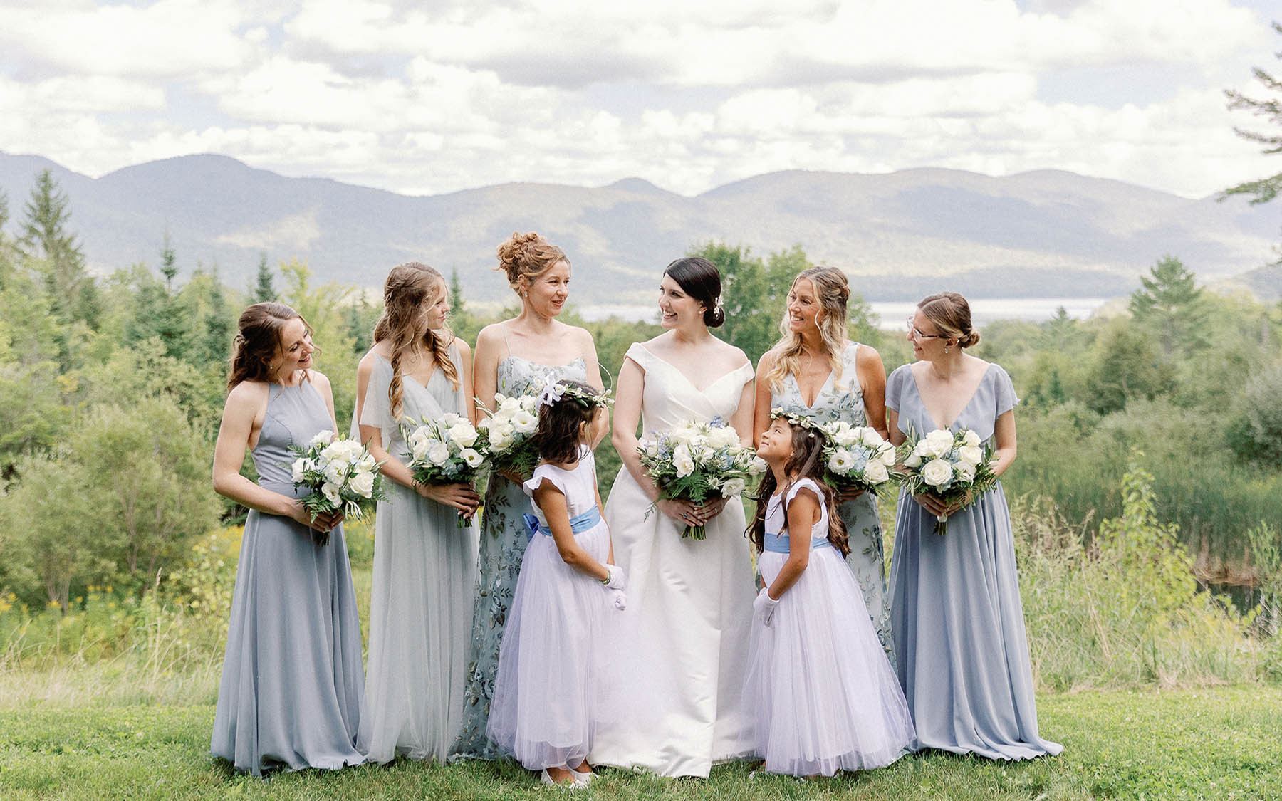 bridal party posing for photo  with woods and blue sky in the background. Bridal party wearing soft blue dresses, bride wearing a traditional white gown.