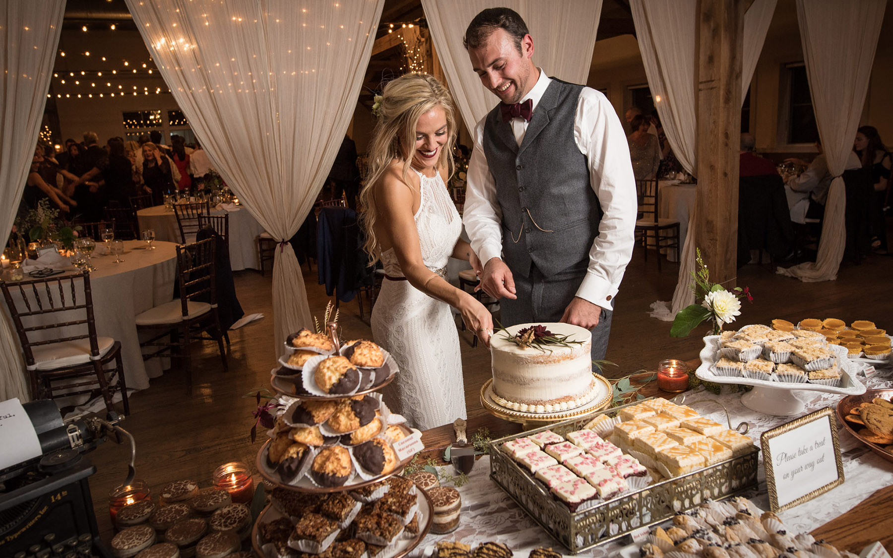 a wedding couple standing in front of a table full of desserts cutting a wedding cake. Drapery and tables visible in the background.