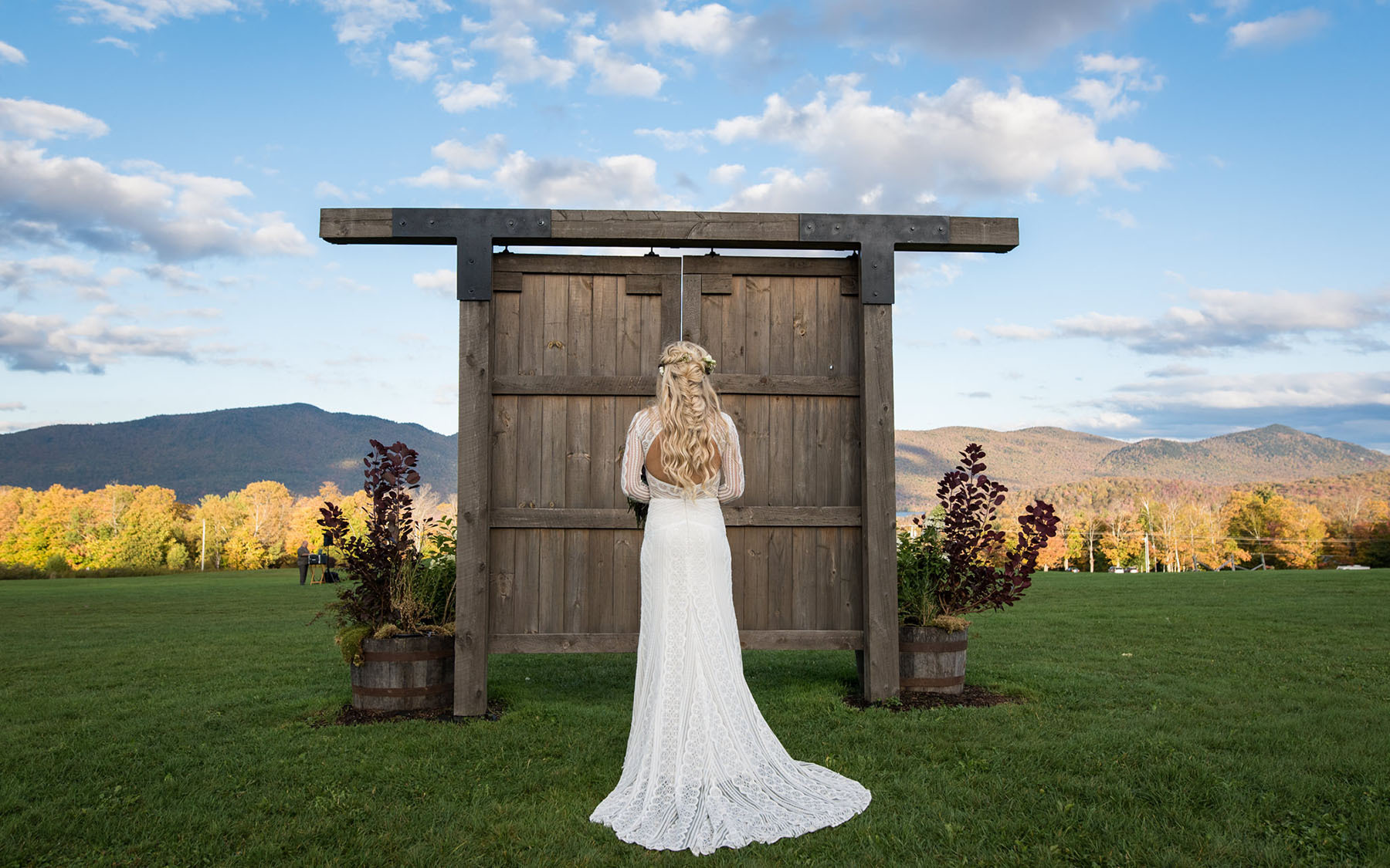 a bride in a white gown, with the trail flowing behind her, standing in front of free standing barn doors in a meadow with the sun setting on the  mountains in the background and blue sky dotted with white clouds.