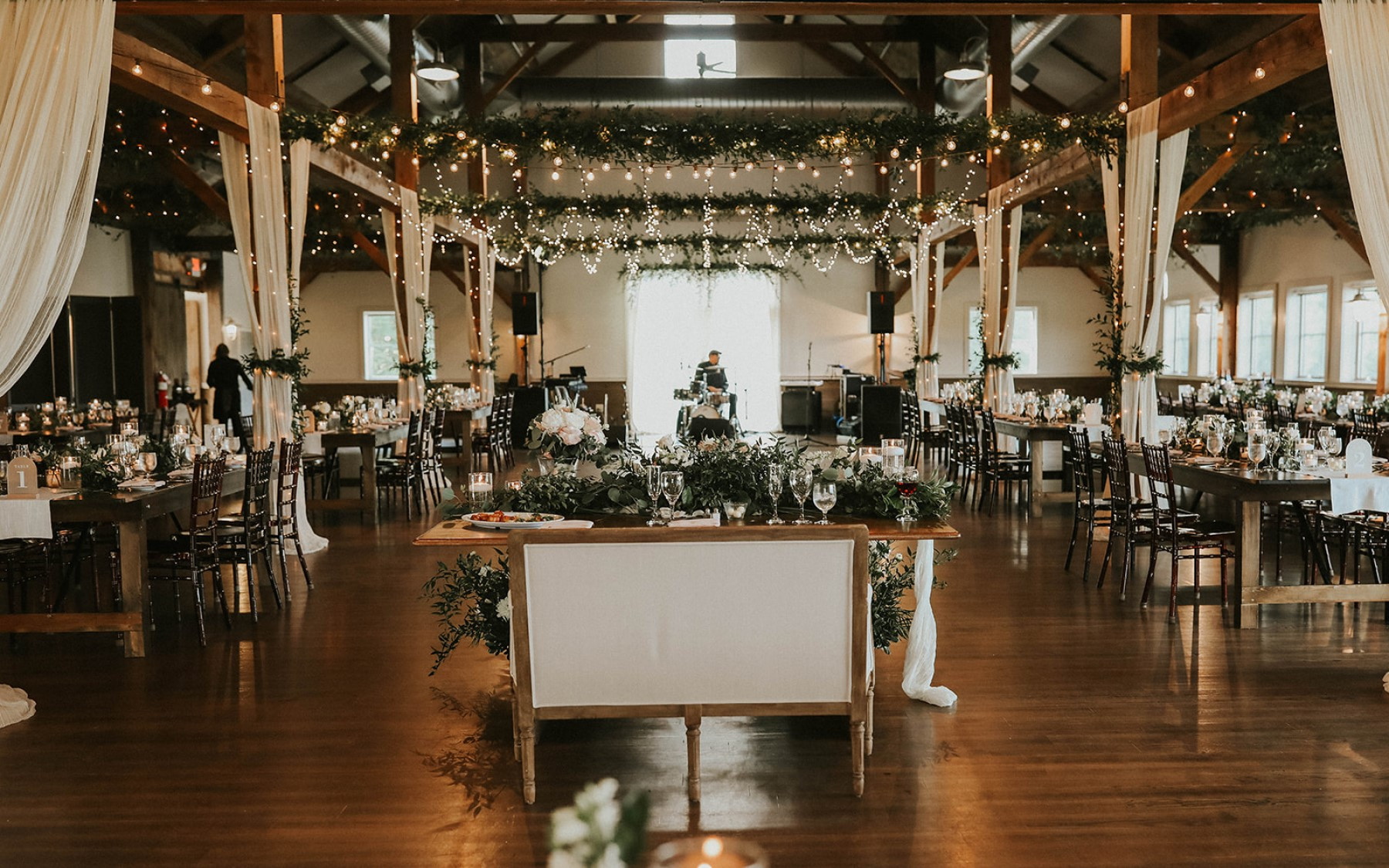 event barn decorated with white florals, drapery, and string lights with sweetheart table in the center with a white loveseat.