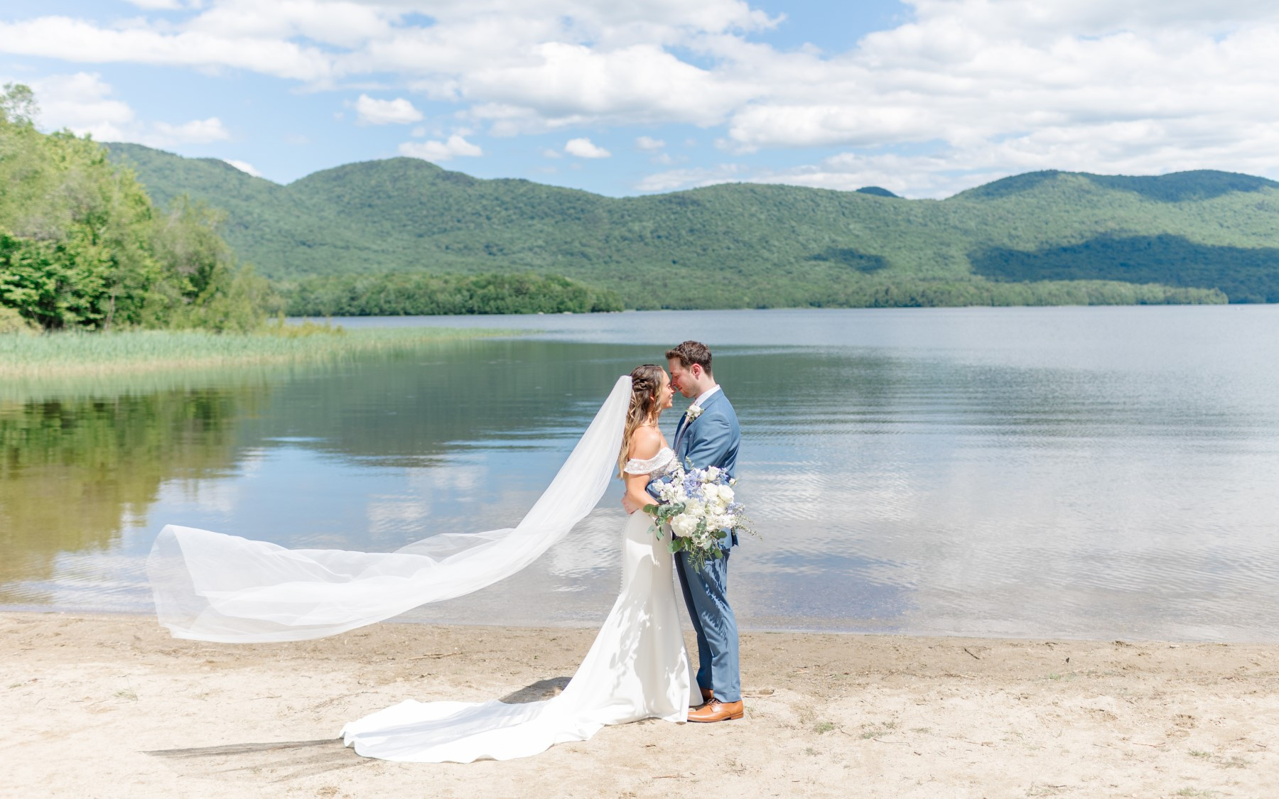 Groom and bride holding each other down by the lake's edge. The brides veil is flowing in the wind and the mountains are in the background.