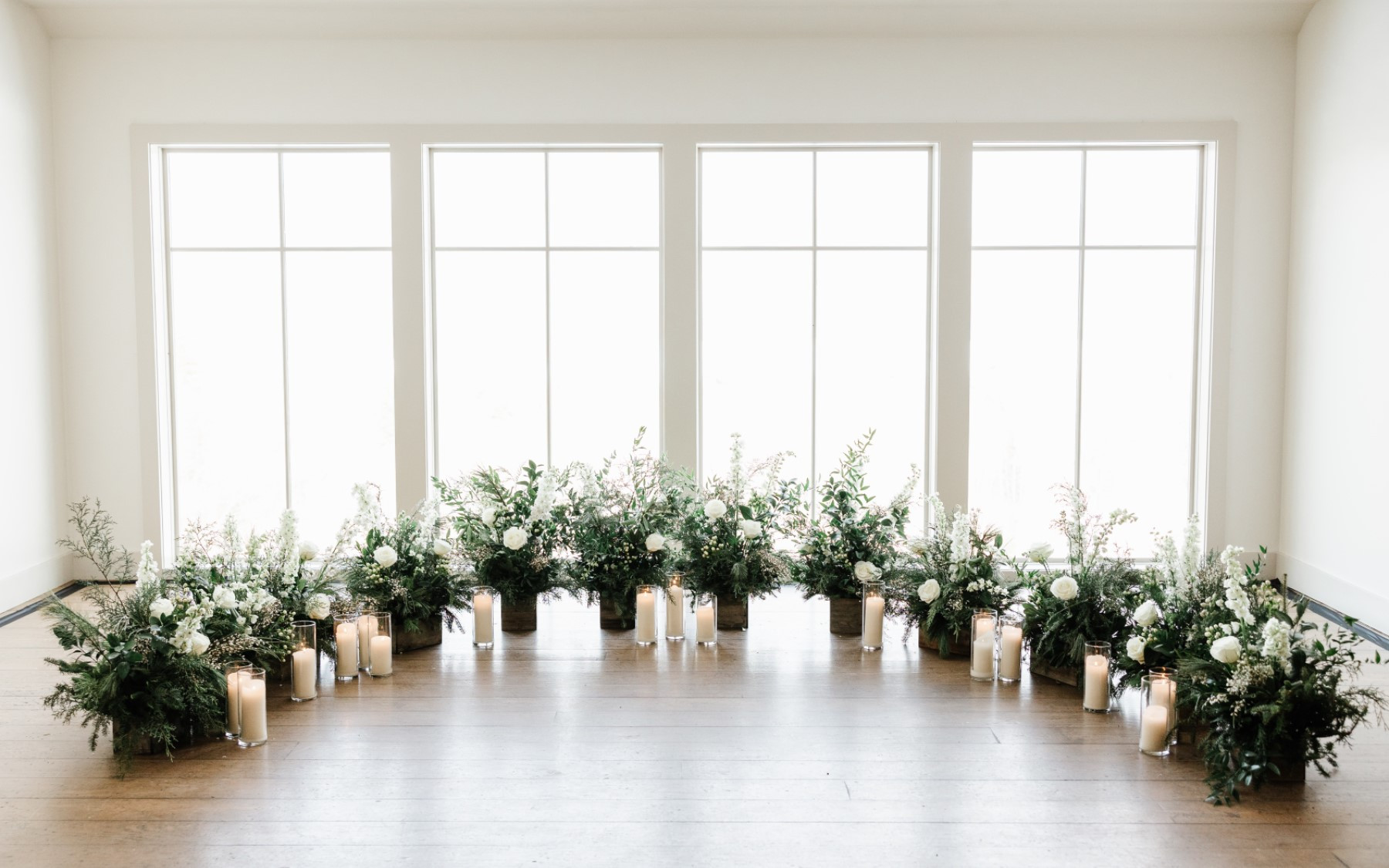 Loft floral design setup featuring white candles in votives, green, white and blue floral arrangements in semi circle with four windows behind.