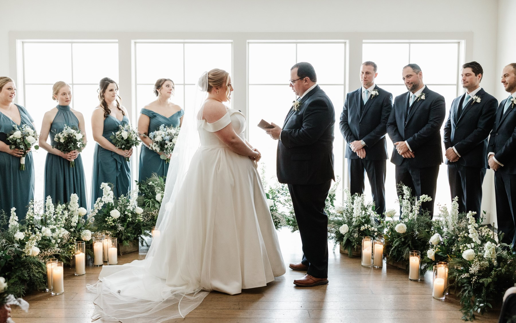 Loft floral ceremony site featuring green/white/blue floral arrangements, with bride and groom in front, and 3 groomsmen and 3 bridesmaids behind them