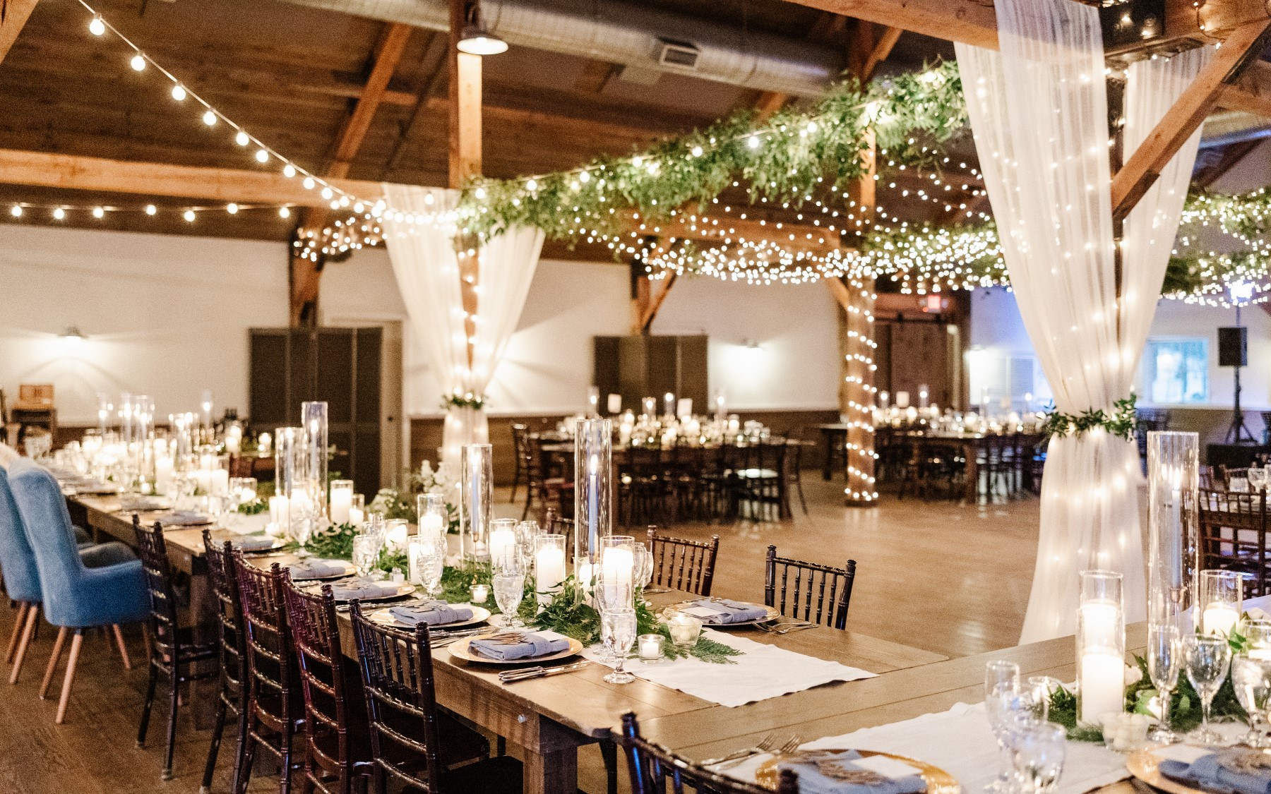 event barn featuring long farmhouse tables, greenery on tables with blue linens, and lights, greenery, and drapery from the top of the event barn.