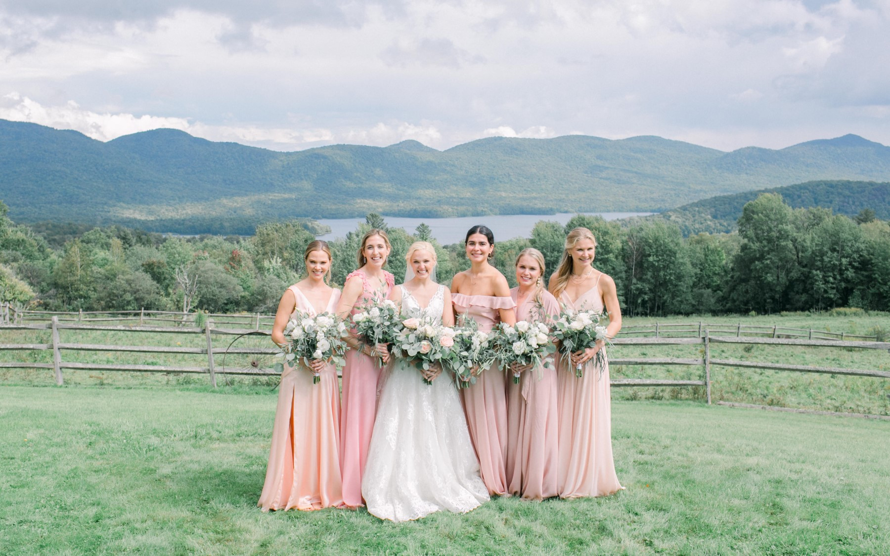 Bride in middle. 5 women in pink dresses, all carrying pink and white bouquets in front of lake and Green Mountains.